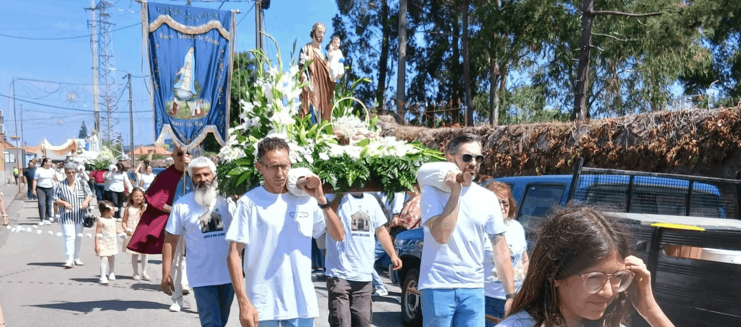 Procissão religiosa ao ar livre com participantes carregando andor de São José com o Menino Jesus, flores brancas e cartaz 'Procissão Solene S. José Rogai por Nós'.