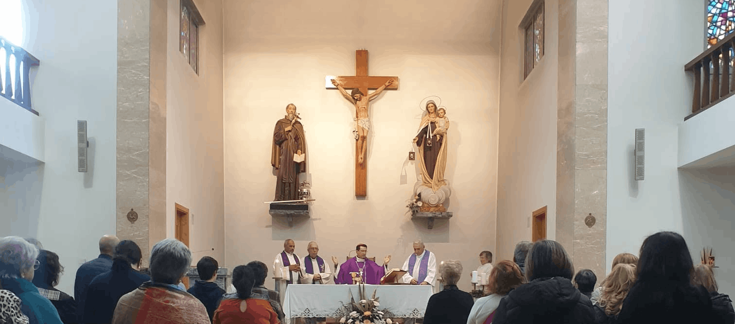 Interior de igreja durante celebração com crucifixo central, imagens de santos, clérigos em vestes roxas e brancas e assembleia voltada para o altar.